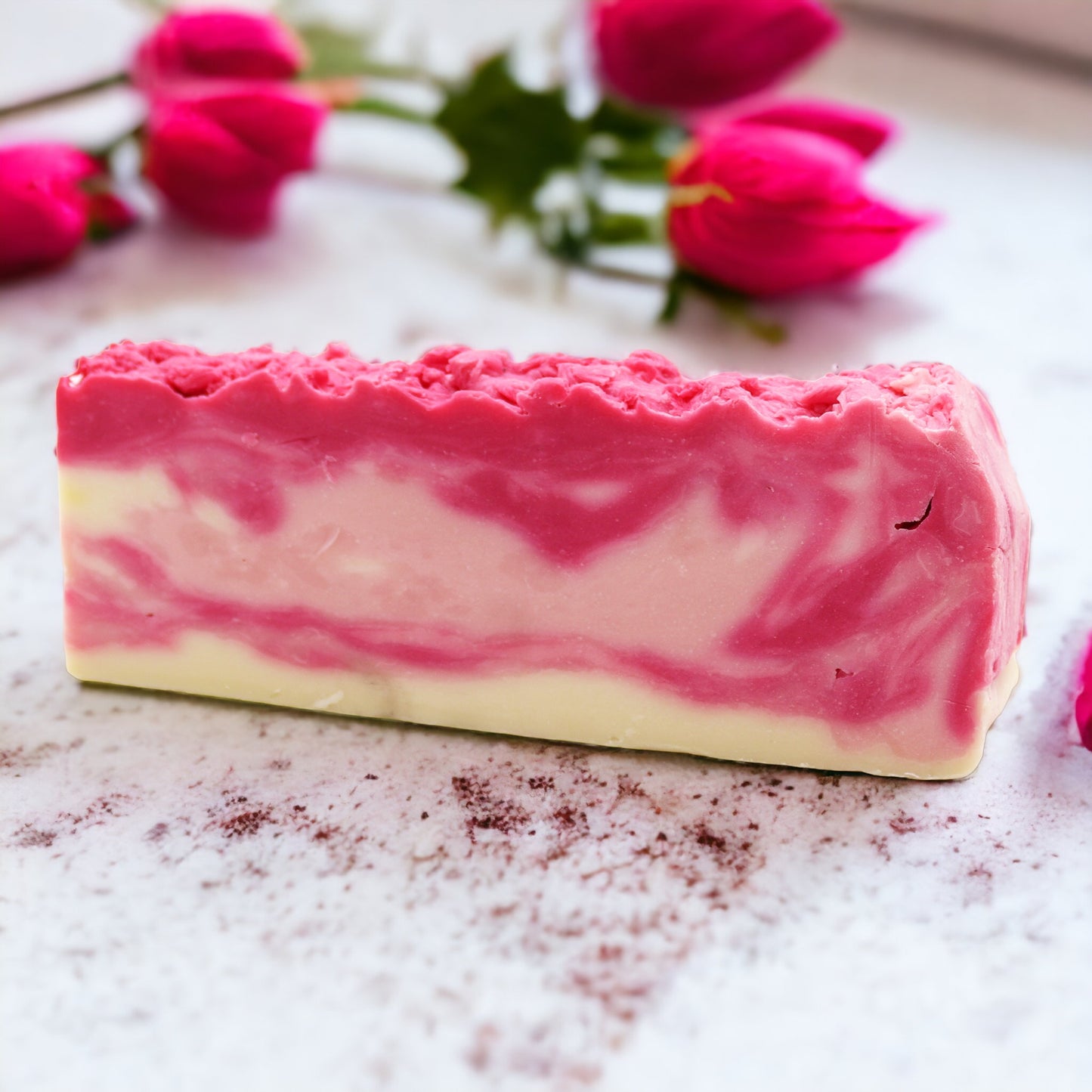 A bar of Rosehip - Olive Oil Soap with a layered design, predominantly pink with a white edge, on a textured grey background with red rose hips and petals.
