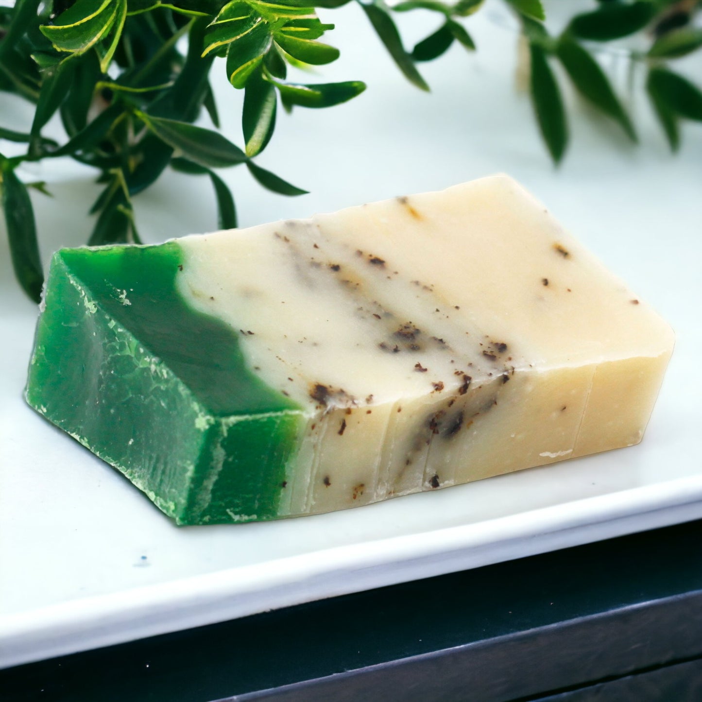 A bar of green tea and olive oil soap with exfoliating particles, placed on a white surface with a plant in the background.