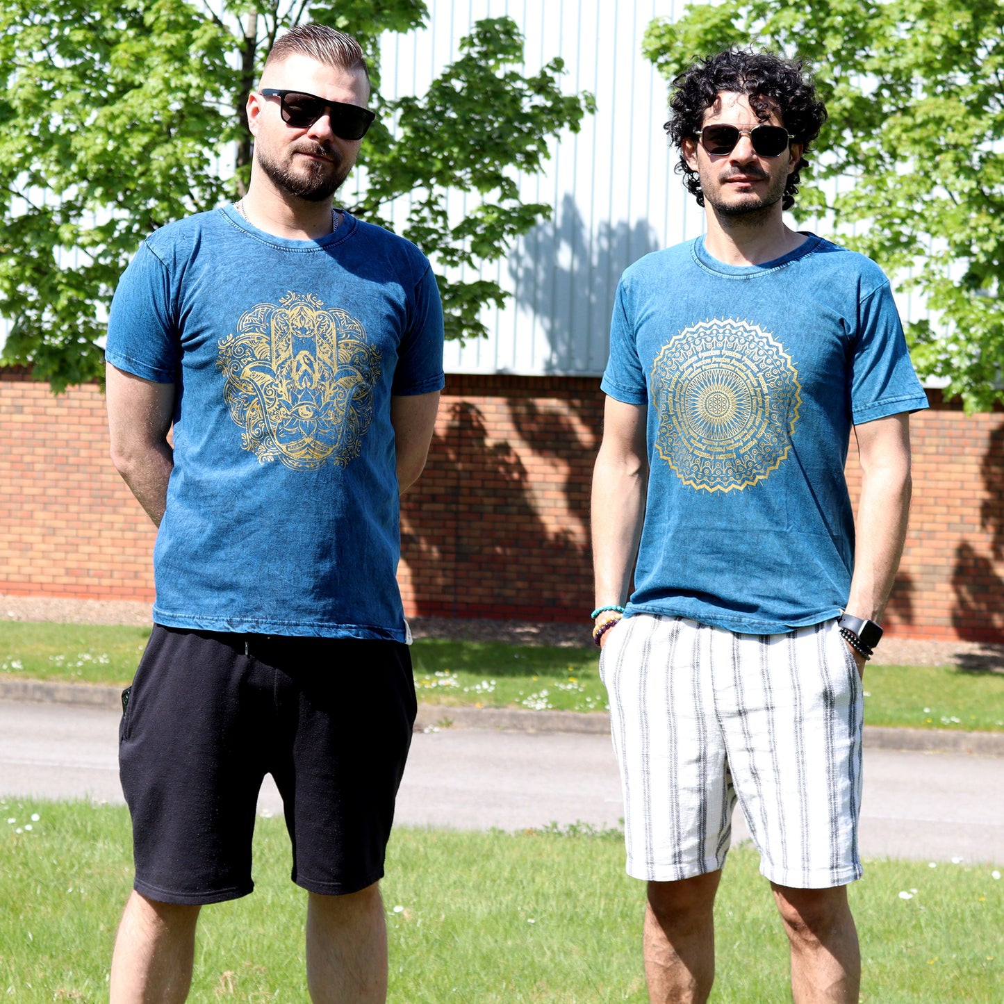Two men wearing blue t-shirts with a circular gold design outdoors.