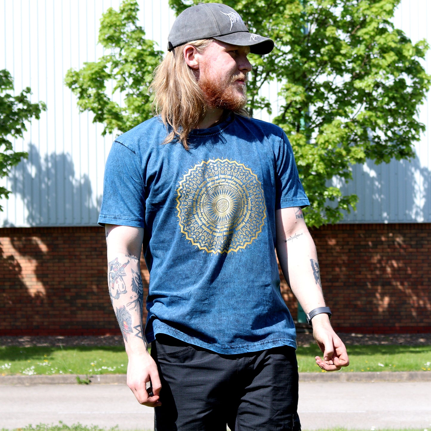 Man with long hair & wearing a blue t-shirt with a yellow mandala design outdoors.