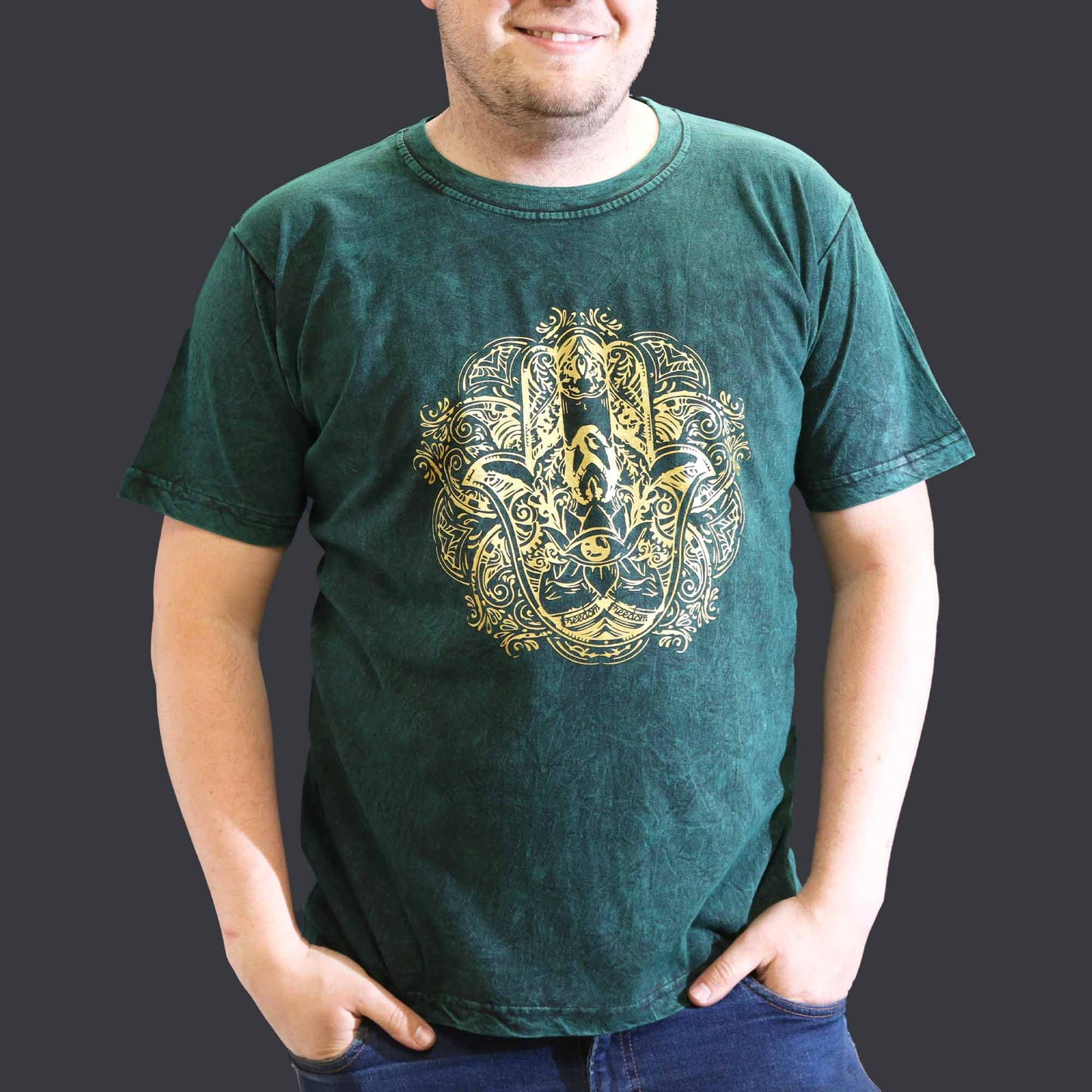 Person with hands in pockets wearing a green t-shirt with a gold Hamsa design on a dark background