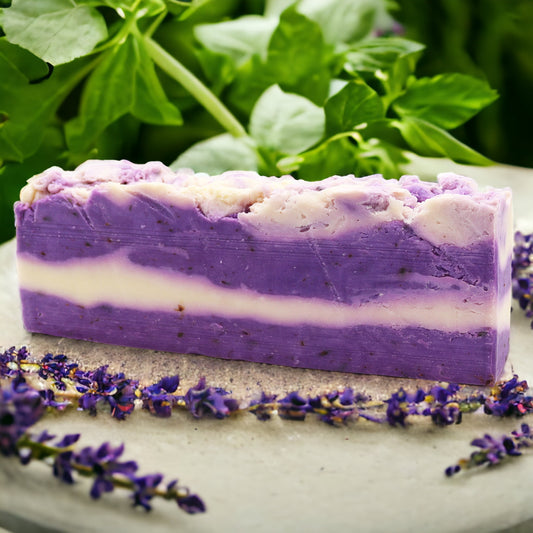 A bar of lavender-colored olive oil soap with white stripes, surrounded by real lavender flowers on a light background.