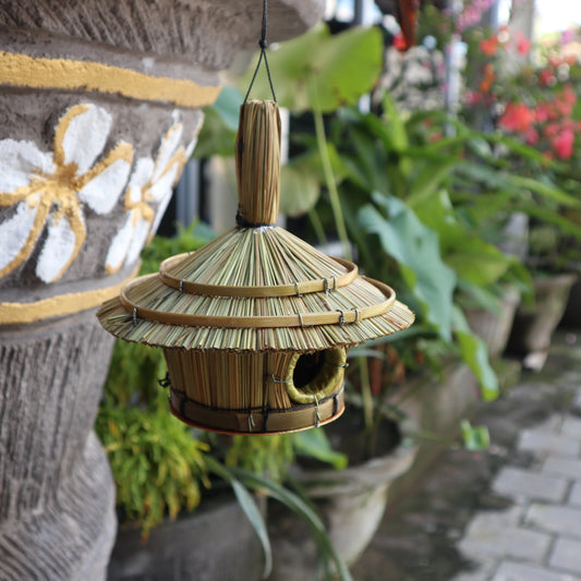 Bamboo birdhouse hanging in a garden setting with plants and a stone wall.