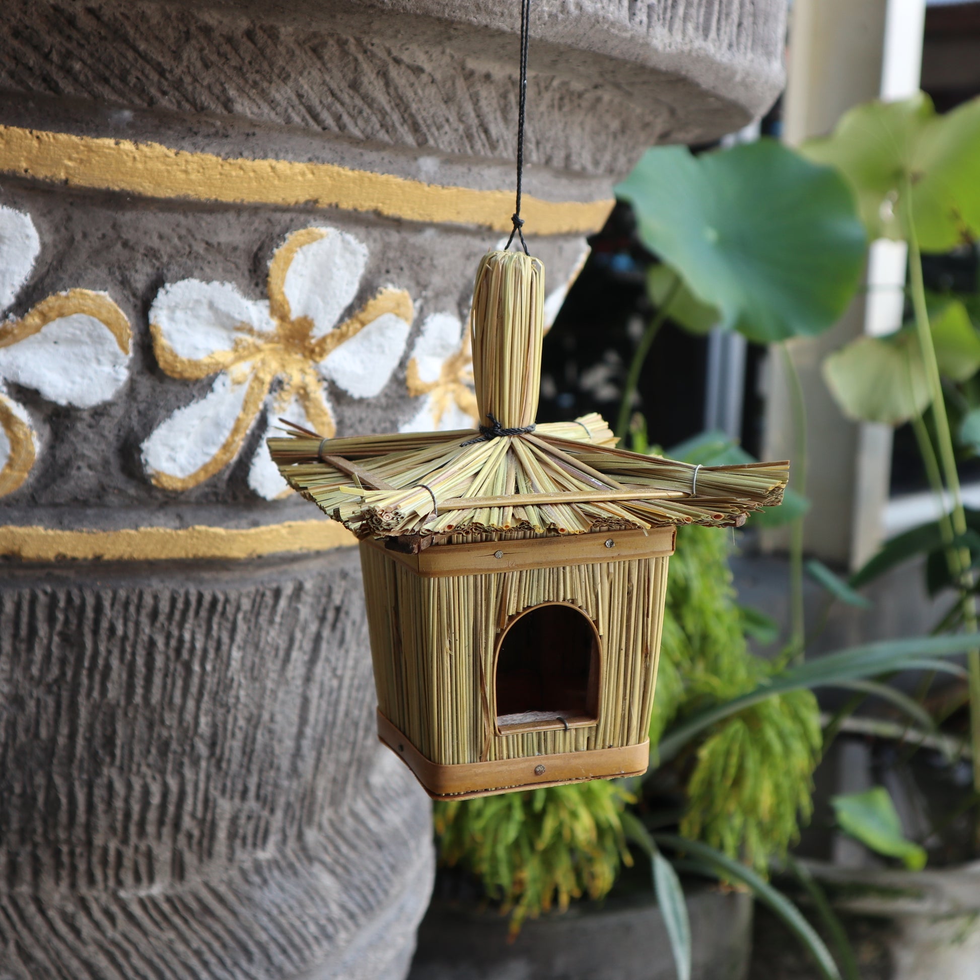 Bamboo birdhouse hanging against a textured wall with plants in the background