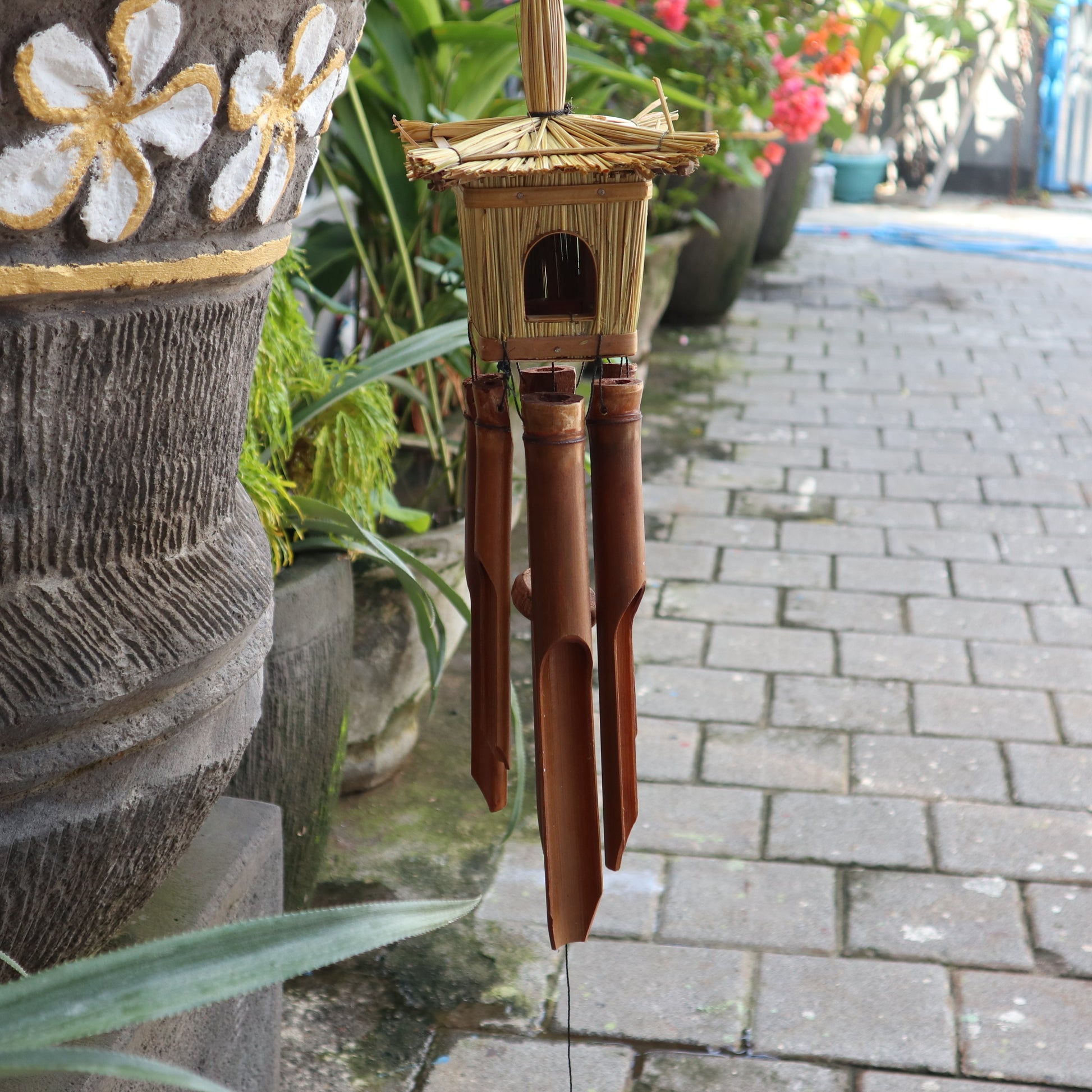 Bamboo wind chime hanging on a stone pillar with plants and a pathway in the background