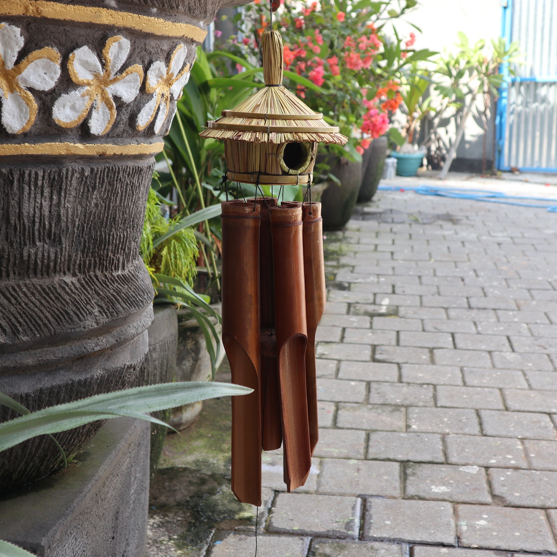 Bamboo wind chime hanging outdoors on a stone pathway with plants and flowers in the background.