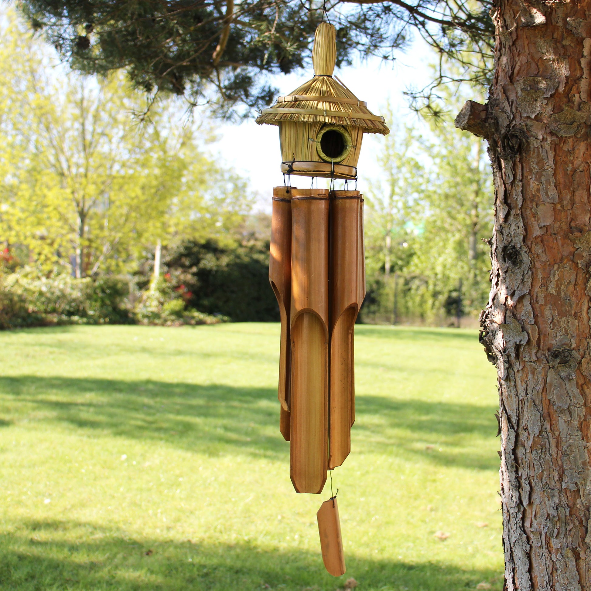 Decorative wind chime with birdhouse attached hanging from a tree in a garden setting.