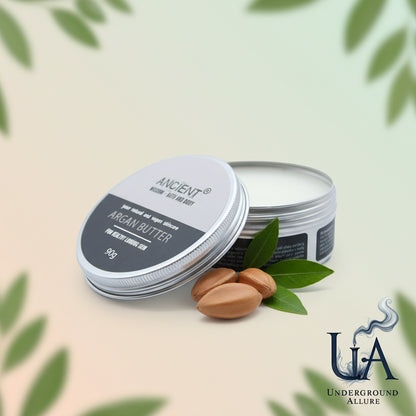 Ancient Wisdom Argan Butter in an open tin, accented by argan nuts and green argan leaves, set against a gentle, wellness-themed background with the Underground Allure logo