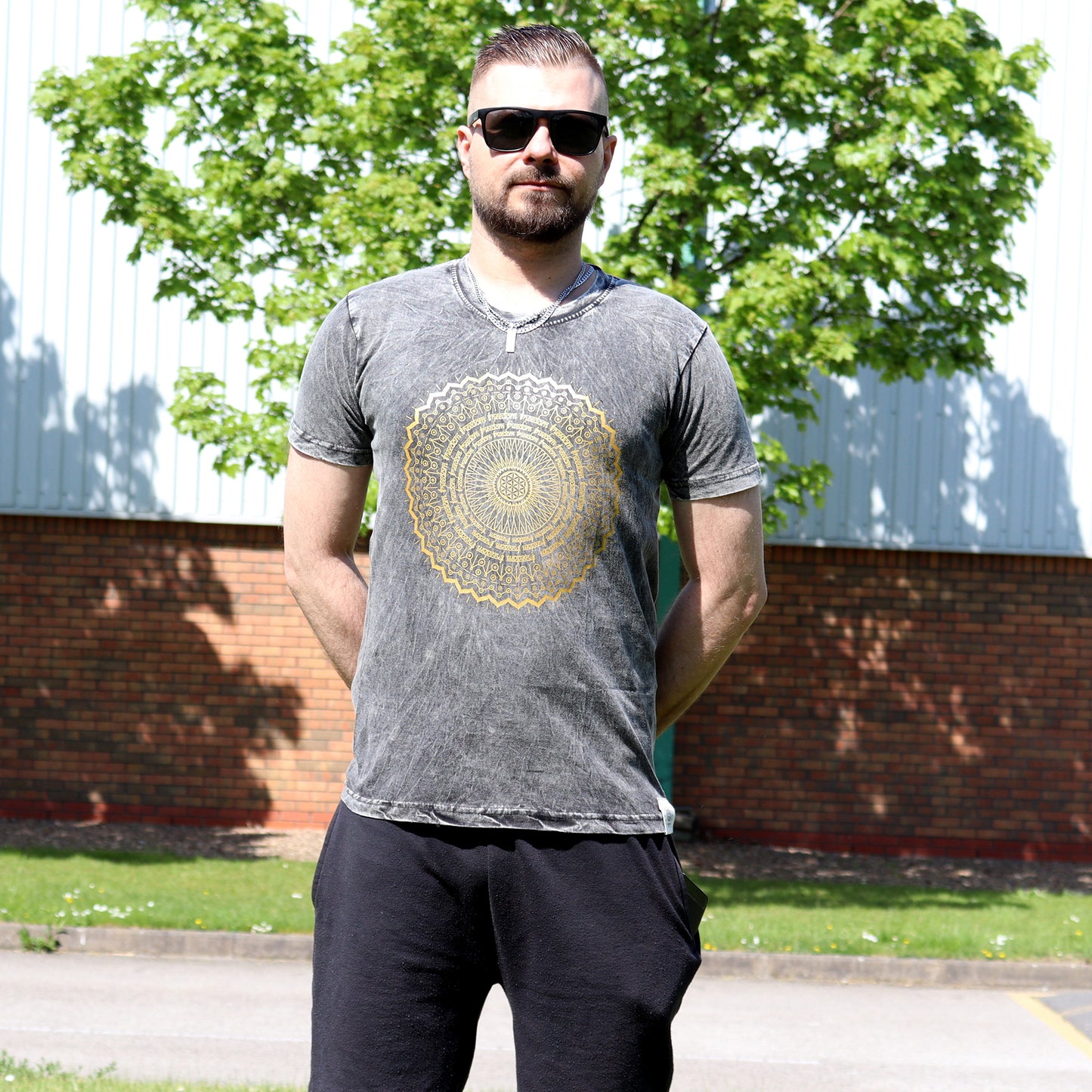 Man wearing  sun glasses & a gray t-shirt with a gold mandala design outdoors.