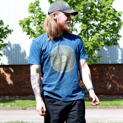 Man with long hair & wearing a blue t-shirt with a yellow mandala design outdoors.