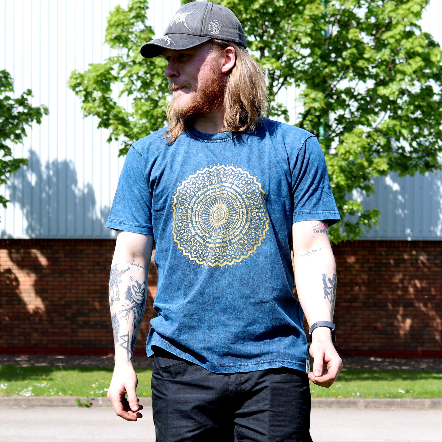 Man with tattoos wearing a blue t-shirt with a yellow mandala design outdoors.