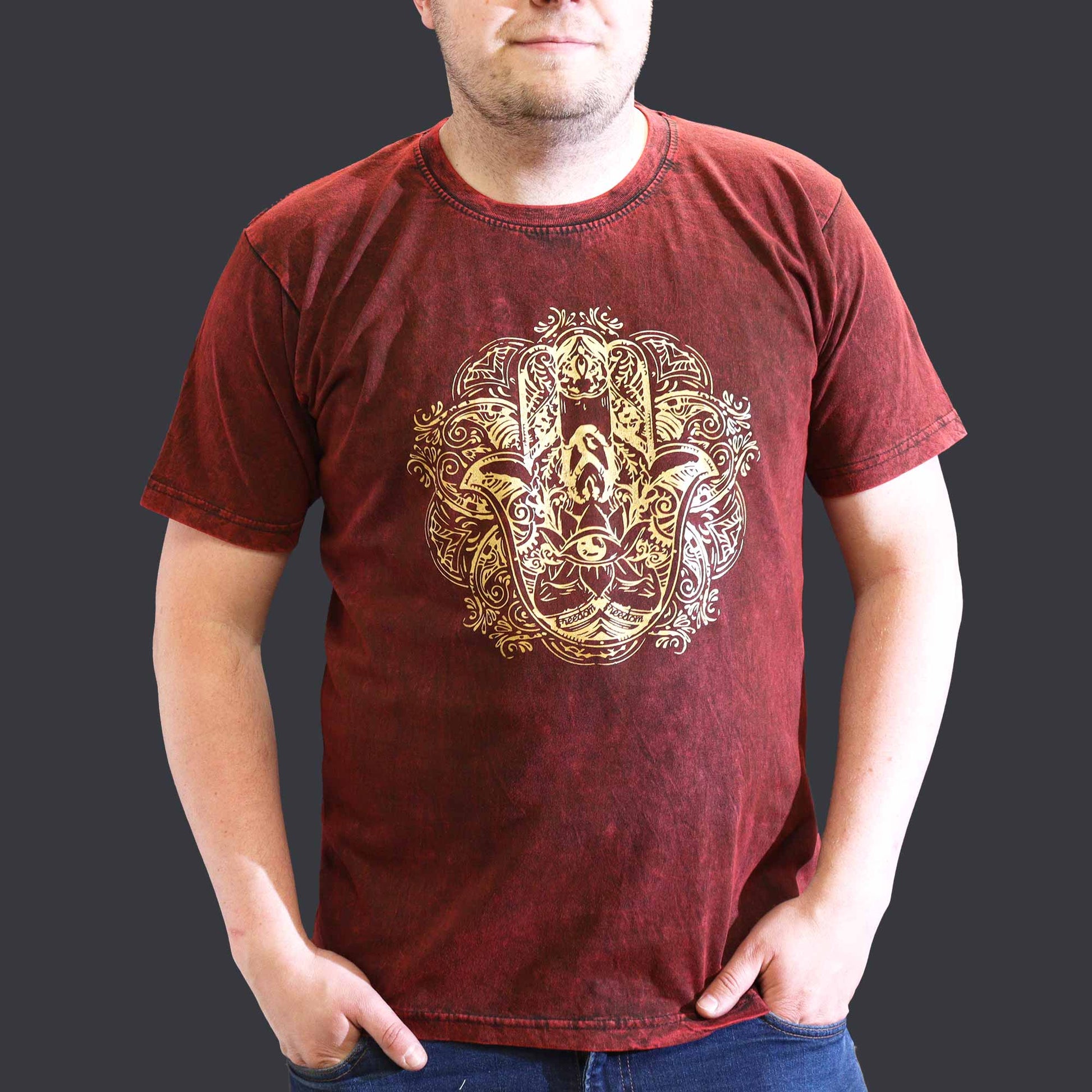 Person with hand in pockets wearing a maroon t-shirt with a Hamsa gold design on a dark background