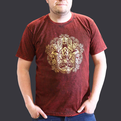 Person with hand in pockets wearing a maroon t-shirt with a Hamsa gold design on a dark background